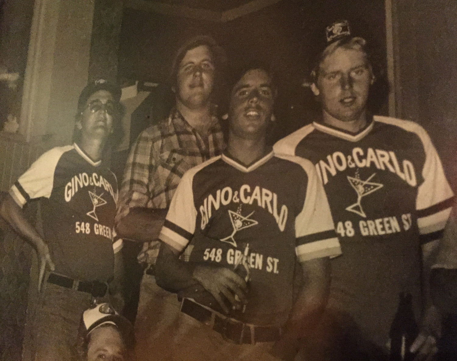 Four men standing together indoors, all wearing matching dark baseball-style jerseys with white sleeves and the text 'GINO & CARLO 548 GREEN ST' plus a martini glass logo.